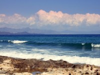 Waves rolling onto a rocky shore under a blue sky, with mountains in the distance