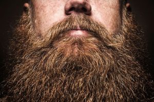 Close-up of a man's lower face with a thick, full reddish-brown beard against a dark background
